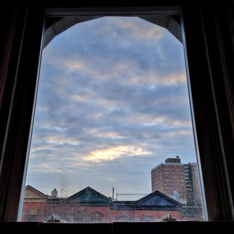 Looking through an arched window seventeen minutes after sunrise the blue sky is filled large bands of chunky light clouds, and there is a patch of sun breaking through in the lower sky. Pointed roofs of Harlem brownstones with red brickwork are across the street, and a taller apartment building can be seen in the distance.