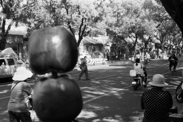 Ilford Pan 100 (FF)

English Alt Text: A black-and-white street scene with people engaged in daily activities. In the foreground, two large fruits on a stick are out of focus. A person in a wide-brimmed hat walks or rides a bicycle on the left. Another person in a patterned shirt and hat is on the right. A third person crosses the street. Bicycles line the sidewalk, and buildings with Chinese signage are in the background.

中文替代文字：
黑白街景，展现人们日常活动。前景中有两颗模糊的大水果串在棍子上。左侧一人戴宽边帽，步行或骑车经过。右侧一人穿花纹衬衫戴帽子。中…