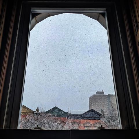 Looking through an arched window seventy-five minutes after sunrise the grey-blue sky is filled with swirling snowflakes. Pointed roofs of Harlem brownstones with red brickwork are across the street, and a taller apartment building can be seen in the distance. The tops of two trees are on the bottom and right, covered in snow. 
