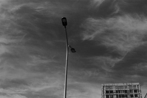 Ilford Pan 100 (FF)

English Alt Text:
A black-and-white photo captures a tall streetlight with two lamps, one higher than the other, standing against a dramatic sky filled with wispy clouds. The streetlight is centered and silhouetted, creating a stark contrast with the textured sky. In the lower right corner, part of a multi-story building with a grid of windows is visible. The building’s rigid geometry contrasts with the organic flow of the clouds. The image evokes a contemplative mood throu…