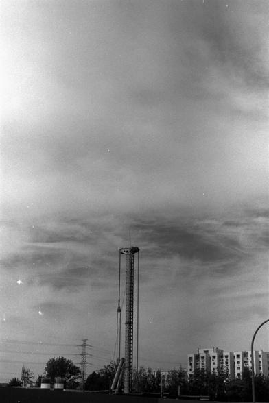 Ilford Pan 100 (FF)

English Alt Text:
A black-and-white photo features a tall construction facility, likely a tower, rising into a cloudy sky. The central tower has cables and a carriage suspended near the top. Below, residential buildings, trees, and electrical transmission towers suggest an urban setting. The tower dominates the composition, emphasizing verticality and scale. The monochrome tone adds a nostalgic, dramatic atmosphere.
中文替代文本：
这张黑白照片展示了一座高耸的建筑设施，可能是塔，直冲云霄。中央塔体配有缆绳，一个舱体悬挂在顶部附近。…