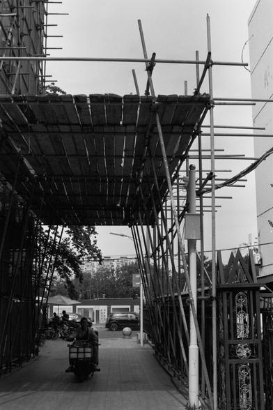 Ilford Pan 100 (FF)

English Alt Text:
A monochrome photo shows a temporary structure made of scaffolding and wooden planks forming a tunnel-like canopy. Metal poles crisscross vertically and diagonally, creating a dense framework. Beneath the structure, a person rides a motorbike carrying cargo. In the background, bicycles are parked, cars are visible, and a decorative gate leads to a building with windows. Trees and distant apartments complete the urban scene. The image highlights daily life …