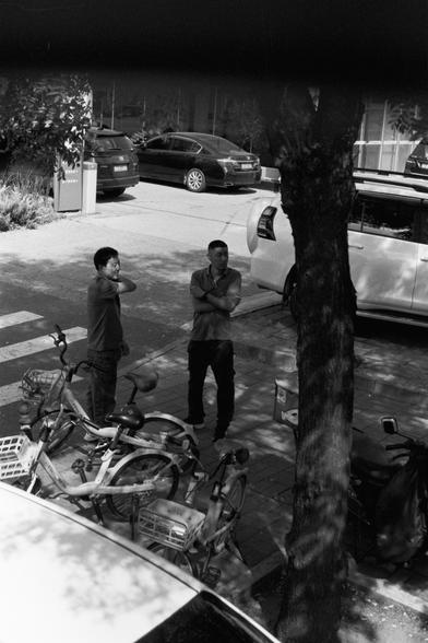 Ilford Pan 100 (FF)

English Alt Text:
A black-and-white street scene shows two people standing on a sidewalk near a tree. One person has an arm raised to their neck, while the other stands with arms crossed. They appear to be conversing or waiting. Several bicycles are parked nearby. In the background, cars are partially visible, including a white vehicle behind the tree and a black sedan. A crosswalk, trash bin, and a building with glass windows complete the urban setting. The lighting and sh…