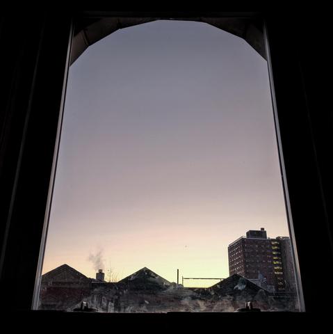 Looking through an arched window fourteen minutes before sunrise the cloudless sky fades from pale yellow at the horizon to light gray up above. Pointed roofs of Harlem brownstones are silhouetted across the street, and a taller apartment building can be seen in the distance. Smoke is coming out of a chimney across the street. The window is grimy on the bottom.