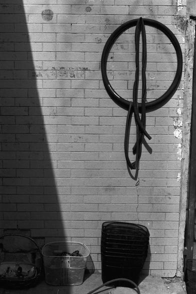 Ilford Pan 100 (FF)

English Alt Text:  
A black-and-white photo of a brick wall with a circular bicycle tire mounted at eye level. Two inner tubes hang loosely through the tire’s center, forming a shape reminiscent of the letter “Q.” Below the tire, wire mesh baskets rest on the ground—one filled with coiled cables or hoses, another stacked with empty baskets. A toolbox or container sits to the left. A diagonal shadow cuts across the left side, adding contrast. The composition is geometric and…