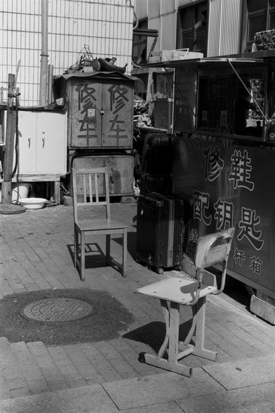 Ilford Pan 100 (FF)

English Alt Text:  
A black-and-white street scene featuring two chairs on a paved sidewalk. One is a simple wooden chair with vertical slats; the other is a worn school chair with a metal frame. Behind them are cabinets and containers with Chinese characters indicating services like car repair, shoe repair, and key duplication. The area is cluttered with wires, suitcases, and tools, suggesting a makeshift repair shop. The setting reflects urban resourcefulness.
中文替代文本：  
这…