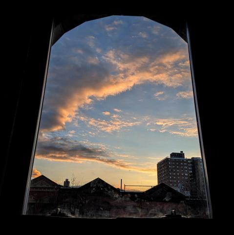 Looking through an arched window fifteen minutes after sunrise the sky fades from yellow at the horizon to baby blue up above. A dramatic arc of gray cloud is lit yellow underneath by the rising sun from behind the houses. Pointed roofs of Harlem brownstones are silhouetted across the street, and a taller apartment building can be seen in the distance.