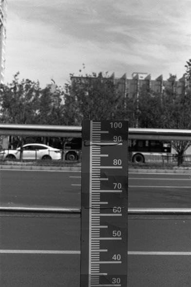 Ilford Pan 100 (FF)

English Alt Text:
A black-and-white image of a vertical measurement gauge beside a road. The gauge is marked from 30 to 100 in increments of 10, with smaller divisions in between. It stands in front of a metal railing. In the background are trees, a white car, a bus, and a multi-story building. The gauge likely measures water levels or clearance height. The urban setting contrasts with the utilitarian design of the gauge.
中文替代文字：  
这是一张黑白照片，展示一条道路旁的垂直测量尺。尺上标有从30到100的数字，每隔10…