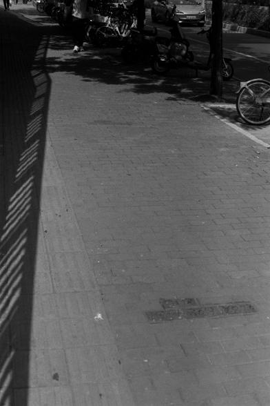 Ilford Pan 100 (FF)

English Alt Text:
A black-and-white street scene showing a sidewalk with shadow patterns cast by a fence or railing. Several scooters and bicycles are parked along the right side. A few pedestrians walk in the distance. A car drives on the road in the background. A metal plate embedded in the sidewalk displays partially legible numbers: "182 303 427." The image captures urban life with strong contrasts and geometric shadows.
中文替代文字：  
这是一张黑白街景照片，画面中人行道上有栏杆投下的阴影图案。右侧停放着几辆电动车…