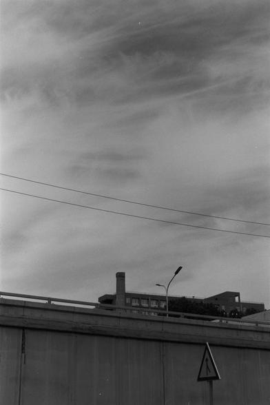 Ilford Pan 100 (FF)

English Alt Text:
A black-and-white photo of a concrete wall in the foreground, with buildings visible above it. One building has a chimney; another is modern with many windows. A streetlight rises above the wall. The sky is filled with wispy clouds and two overhead utility wires stretch across the image. In the bottom right corner, a triangular traffic sign with an upward arrow indicates a one-way direction. The image contrasts industrial and residential elements with dram…