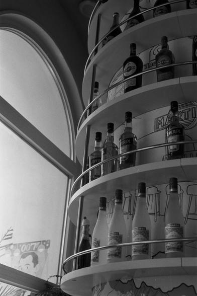 Ilford Pan 100 (FF)

English Alt Text:
A black-and-white photo of a multi-tiered circular shelf filled with bottles of alcoholic beverages. Brands like Martini, Aperol, and Luxardo Limoncello are visible. The shelf has metal railings and is part of a modern interior space. Behind it, a wall features illustrated graphics of bar stools and signage reading "Martini." To the left, a large arched window lets in natural light. Outside the window, a poster with a stylized face and the text "007" is pa…
