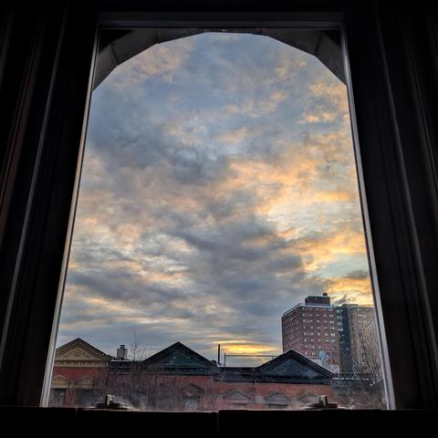 Looking through an arched window twenty-three minutes after sunrise the blue sky is filled with complex gray and white clouds, with a few patches of sun breaking through. Pointed roofs of Harlem brownstones with red brickwork are across the street, and a taller apartment building can be seen in the distance. 