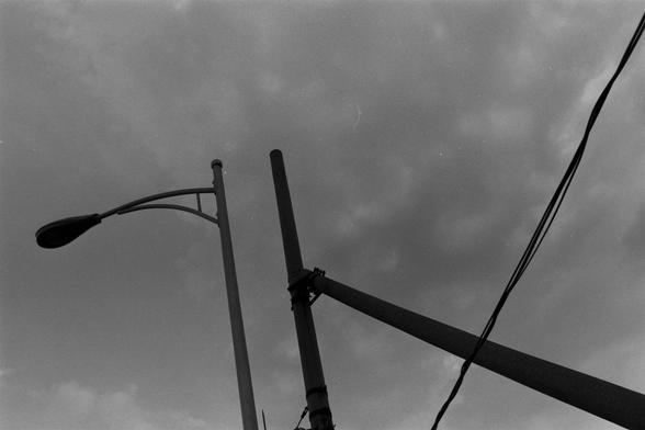 Ilford Pan 100 (FF)

English Alt Text:
A black-and-white photograph shows a streetlight pole and a utility pole with multiple cables stretching across the frame. The streetlight has a curved arm extending leftward with a lamp at the end. The utility pole includes a diagonal support beam. Overhead wires crisscross the cloudy sky, forming geometric patterns. The moody sky and intersecting lines create a visually striking and structured composition.
中文替代文本：  
这是一张黑白照片，画面中有一根街灯杆和一根电线杆，多条电缆横跨画面。街灯杆向…