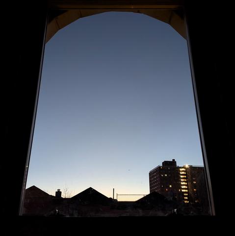 Looking through an arched window thirty-eight minutes before sunrise the cloudless sky fades from white at the horizon to skate blue up above. Pointed roofs of Harlem brownstones are silhouetted across the street, and a taller apartment building can be seen in the distance.