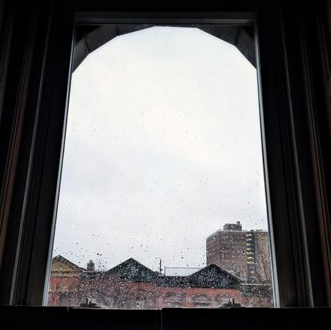 Looking through an arched window forty minutes after sunrise the sky is full of gray rain clouds and the window is covered in rain drops. Pointed roofs of Harlem brownstoneswuth red brickwork are across the street, and a taller apartment building can be seen in the distance.
