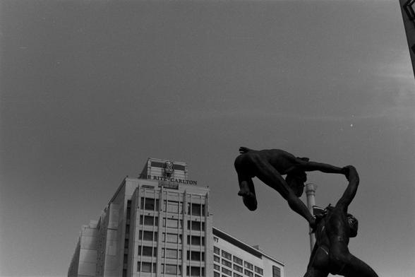 Ilford Pan 100 (FF)

English Alt Text: A black-and-white photograph captures a dramatic bronze sculpture of two human figures in motion. One figure stands firmly on the ground, arms raised, appearing to lift or support the second figure, who is airborne in a dynamic leap. Their bodies are muscular and expressive, frozen in a moment of intense physical interaction. The sculpture evokes themes of strength, trust, and elevation. Behind the sculpture, a tall modern building with the sign “THE RITZ-…
