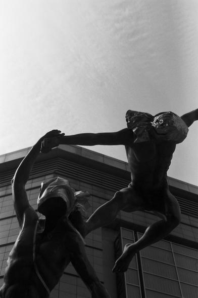 Ilford Pan 100 (FF)

English Alt Text: A monochrome photograph showcases a surreal sculpture of two male figures in motion. One figure is airborne, leaping above the other, who stands below with arms extended. Both figures have their faces obscured by cloth or metallic material, adding mystery and anonymity. Their bodies are muscular and stylized, frozen in a moment of dynamic interaction. The sculpture suggests themes of transformation, struggle, or transcendence. In the background, a modern b…
