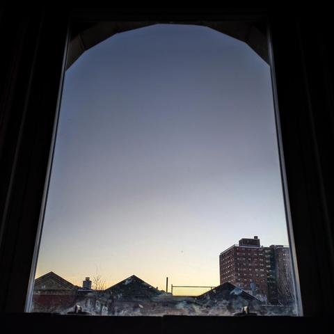 Looking through an arched window at the moment of sunrise the cloudless sky fades from pale yellow at the horizon to dusty blue above. Pointed roofs of Harlem brownstones are silhouetted across the street, and a taller apartment building can be seen in the distance. The window is grimy on the bottom.