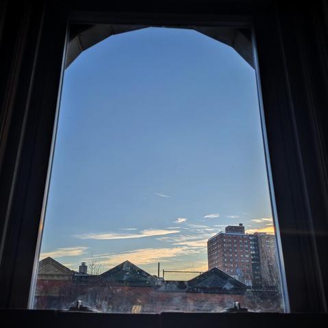 Looking through an arched window forty-nine minutes after sunrise the blue sky is marked with a few scattered skinny white and yellow clouds above the hotizon. Pointed roofs of Harlem brownstones are silhouetted across the street, and a taller apartment building can be seen in the distance. The window is grimy on the bottom.