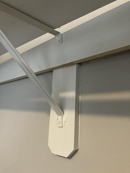 A picture of a vertical piece of wood mounted tot he wall, with a horizontal wood piece acting as a rail. Critically, there is a gap between the horizontal wood piece and the wall the depth of the vertical wood piece because it’s been installed, not to the wall, but atop the vertical wood piece. There is part of the metal support bracket in view. It’s bent because the attachment points are not level. Dumb. 