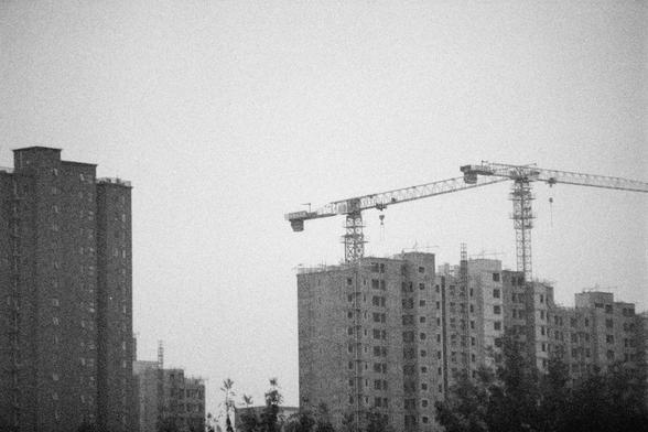 Ilford HP5 Plus 400 (FF)

English Alt Text:  
A monochrome cityscape captures several high-rise apartment buildings under construction. Two large tower cranes dominate the skyline, indicating active development. The buildings vary in completion, with some showing exposed concrete and window frames. In the foreground, dark silhouettes of trees add depth and contrast. The image conveys a sense of urban expansion and industrial progress, with a gritty, documentary-style tone.
中文替代文字：  
这是一幅黑白城市景观图…