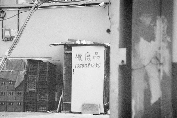 Ilford HP5 Plus 400 (FF)

English Alt Text:  
A gritty black and white photo depicts an alley or back area with stacked plastic crates and a small structure featuring a white door. Above the door, pipes and wires run along the wall. On the door, handwritten Chinese characters read “收废品” (collecting recyclables) followed by a phone number. The scene suggests a local recycling business or informal service, with a utilitarian and raw aesthetic.
中文替代文字：  
这是一张黑白照片，画面为一条后巷或角落，堆放着塑料箱子，旁边是一间小屋，屋门为白色。门…