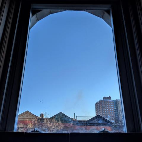 Looking through an arched window ninety minutes after sunrise the blue sky is cloudless. Pointed roofs of Harlem brownstones with red brickwork are across the street, and a taller apartment building can be seen in the distance. The leafless tops of two trees are on the bottom and right. The window is grimy on the bottom.