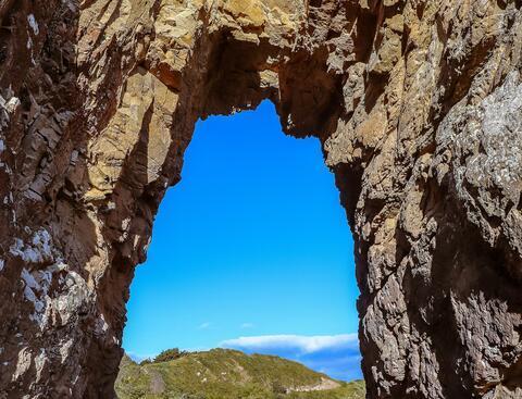 A view through a craggy rock showing a hill against a blue sky.