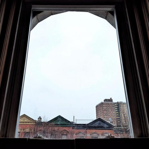 Looking through an arched window twenty-six minutes after sunrise the sky is white and cloudless. Pointed roofs of Harlem brownstones with red brickwork are across the street, and a taller apartment building can be seen in the distance. The leafless tops of two trees are on the bottom and right.