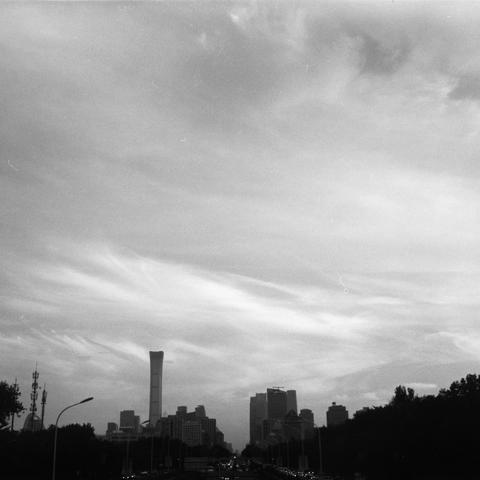 Lucky SHD 400 (6x6)

English Alt Text:
A black-and-white photograph captures a modern cityscape from street level. A wide road stretches into the distance, flanked by evenly spaced trees and streetlights. Vehicles are visible on the road, suggesting light traffic. In the background, a cluster of high-rise buildings dominates the skyline, with one uniquely shaped skyscraper standing prominently at the center. The sky is filled with wispy, textured clouds, adding depth and atmosphere. The monochr…