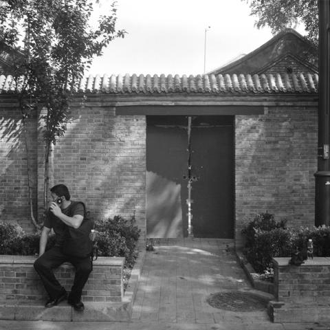 Lucky SHD 400 (6x6)

English Alt Text:
A monochrome photo shows a man sitting on a low brick wall in front of a traditional East Asian building. He wears dark clothes and a backpack, holding a phone to his face as if talking. Behind him is a brick structure with a tiled roof and a large, weathered double door. Small bushes and trees frame the walkway leading to the door. A bottle and other items rest on the wall nearby. The scene blends modern activity with historical architecture.

中文替代文字：
这是一…