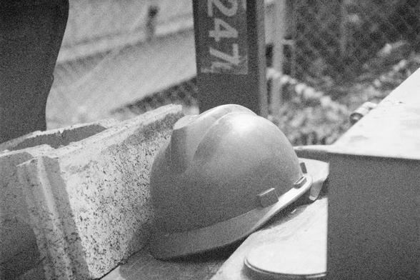 Ilford HP5 Plus 400 (FF)

English Alt Text:
A gritty black-and-white photo of a construction site. A hard hat rests beside a concrete cinder block on a rough surface. Behind them, a chain-link fence and a vertical post marked with the number "247" suggest an industrial setting. The image evokes themes of manual labor, safety, and the physicality of construction work.
中文替代文字：
这是一张黑白照片，展现了一个施工现场。一顶安全帽放在粗糙的地面上，旁边是一个混凝土空心砖。背景中有铁丝网围栏和一根标有“247”数字的垂直柱子，暗示这是一个工业环境。画面传达了体力劳动、安全防护和建筑工作的真实感。