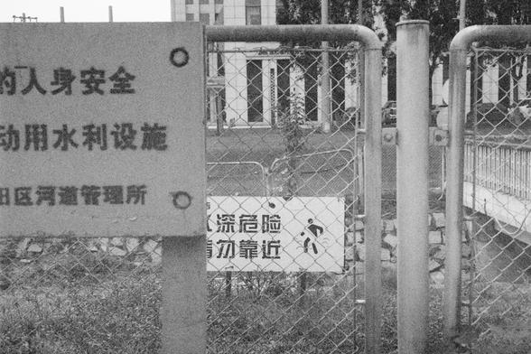 Ilford HP5 Plus 400 (FF)

English Alt Text:
A black-and-white image of a fenced-off area near a waterway. Metal gates and fencing block access to a facility behind, with warning signs in Chinese. One sign warns of deep water danger and advises people to stay away. A bridge and building are visible in the background. The scene emphasizes restricted access and public safety.
中文替代文字：
这是一张黑白照片，显示一个靠近水道的封闭区域。金属栅栏和大门阻止进入，上面挂有中文警示牌。其中一块牌子警告“深水危险，请勿靠近”。背景中可见桥梁和建筑物。画面强调了限制进入和公众安全的重要性。