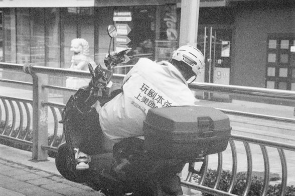 Ilford HP5 Plus 400 (FF)

English Alt Text:
A black-and-white photo of a person riding a scooter or motorcycle on a sidewalk. The rider wears a helmet and a jacket with Chinese text promoting Meituan services. A metal railing lines the path. In the background, a building with glass windows, a stone lion statue, and directional signs are visible. The image blends urban delivery culture with traditional architecture.
中文替代文字：
这是一张黑白照片，画面中一人骑着电动车或摩托车行驶在人行道上，身穿印有中文广告语的外套，宣传美团服务。人行道旁有金属栏杆。背景中可见玻璃窗建筑、…