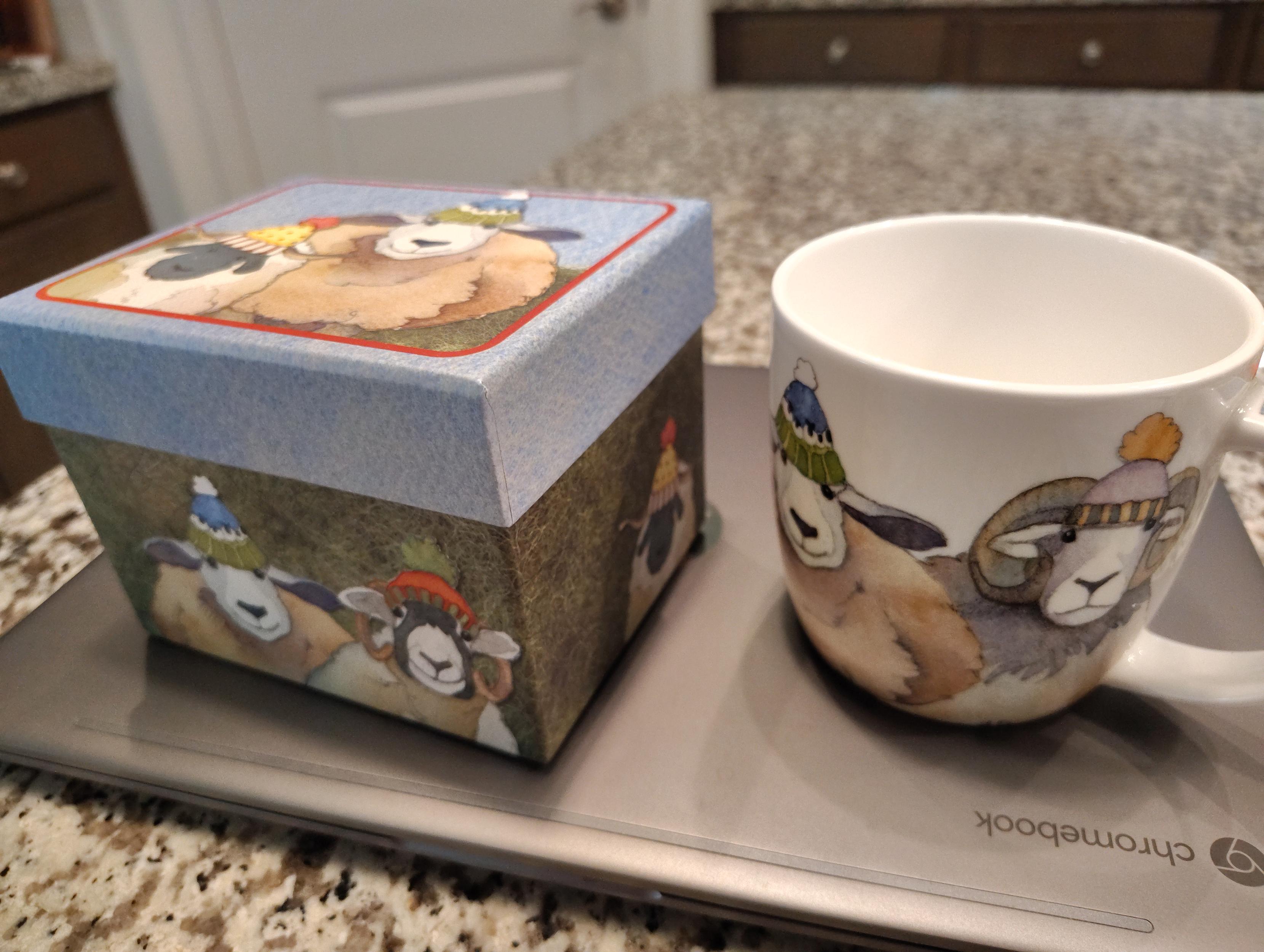 A large white coffee mug is shown next to a decorative box it came in. The mug and the box have illustrations of chubby wooly sheep, each wearing a colorful wool cap with a big pom pom on top. All of the sheep, who are different shades of white, tan, and grey, are smiling