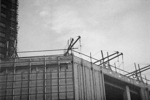 Ilford HP5 Plus 400 (FF)

English Alt Text:
A grainy black-and-white photo of a construction site. In the foreground, a partially built concrete structure features scaffolding and hoisting equipment with cables and pulleys. On the left, a taller building is covered in scaffolding and safety netting. The image captures the industrial atmosphere of urban development, with strong vertical and diagonal lines. The monochrome tone adds a documentary feel, emphasizing the raw materials and mechanical …
