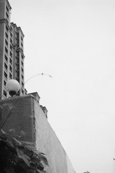 Ilford HP5 Plus 400 (FF)

English Alt Text:
A black-and-white photo of a tall, multi-story building with rows of rectangular windows and ornate architectural detailing. In the foreground, a concrete wall supports a spherical streetlamp mounted on a short pole. Sparse foliage appears in the bottom left corner. Above the building, a faint arc—possibly a wire or cable—stretches across the sky, with small objects attached. The sky is blank and overcast, creating a stark contrast with the building’s…
