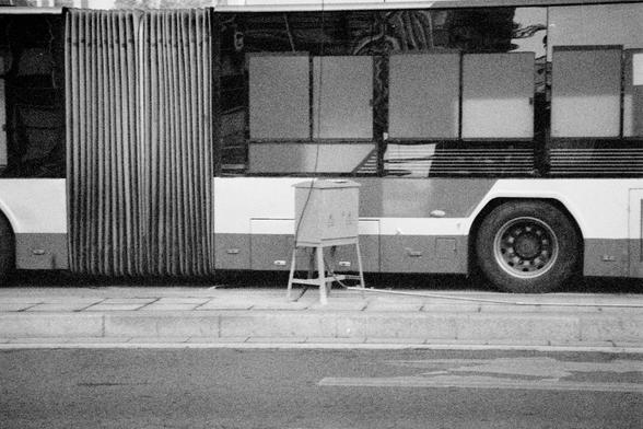 Ilford HP5 Plus 400 (FF)

English Alt Text:
A black-and-white photo of an articulated city bus parked at a curb. The bus features large windows and an accordion-style joint in the middle. In front of the bus, a small utility box stands on four legs, connected to the bus by a cable. The sidewalk and curb are visible, suggesting an urban setting. The image focuses on public transportation infrastructure, with a minimalist composition and subdued tones.
中文替代文字：
这是一张黑白照片，展示了一辆停靠在路边的城市铰接式公交车。公交车中部有风…