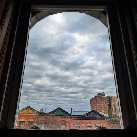 Looking through an arched window sixty-eight minutes after sunrise the sky is filled with chunky gray winter clouds. Pointed roofs of Harlem brownstones with red brickwork are across the street, and a taller apartment building can be seen in the distance. The leafless tops of two trees are on the bottom and right.