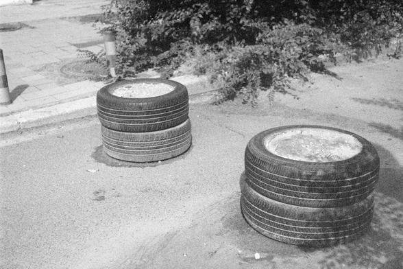 Ilford HP5 Plus 400 (FF)

English Alt Text:
Two stacks of car tires are placed on a paved road near a sidewalk. Each stack consists of three tires, with the top tire filled with concrete or a similar material, possibly serving as a barrier or planter. The tires are worn and dark, contrasting with the lighter pavement. Behind them is a sidewalk made of rectangular paving stones, a curb, and several bollards with striped markings. Dense foliage from nearby bushes or trees fills the background. Th…