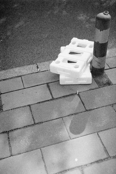 Ilford HP5 Plus 400 (FF)

Two stacked white plastic objects, likely Styrofoam packaging materials, sit on a tiled sidewalk. Next to them is a black and white striped bollard, used to prevent vehicle access. A small cylindrical object lies nearby on the ground. The sidewalk is made of rectangular paving stones. The image is in black and white, highlighting the contrast between the clean, geometric sidewalk and the discarded materials. The composition draws attention to urban textures and the pre…