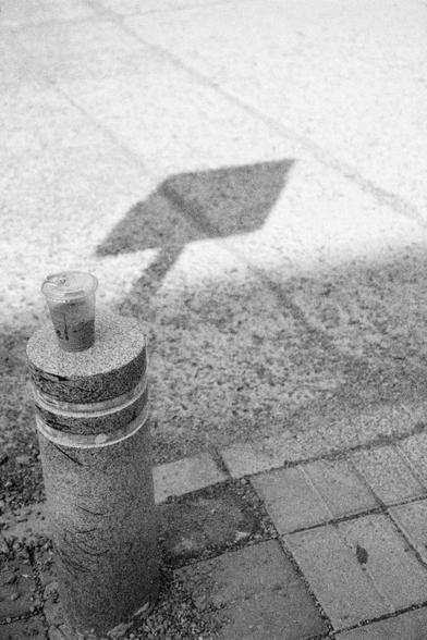 Ilford HP5 Plus 400 (FF)

English Alt Text:
A cylindrical stone bollard stands on a tiled sidewalk. On top of it sits a plastic cup with a lid, likely containing a beverage. The cup is centered and casts a faint shadow. In the background, a sharp diamond-shaped shadow of a traffic or directional sign is cast on the ground, though the actual sign is not visible. The sidewalk is made of square tiles, and the image is in black and white, creating a dramatic interplay of light and shadow. The compo…