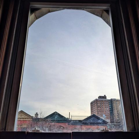 Looking through an arched window two hours after sunrise the blue-gray sky is filled with murky clouds. Pointed roofs of Harlem brownstones with red brickwork are across the street, and a taller apartment building can be seen in the distance. The leafless tops of two trees are on the bottom and right. The window is grimy on the bottom.