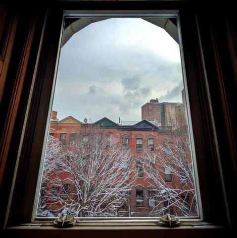 Looking through an arched window two hours after sunrise the sky is filled with winter clouds. Pointed roofs of Harlem brownstones with red brickwork are across the street, and a taller apartment building can be seen in the distance. Several leafless trees are covered in snow.