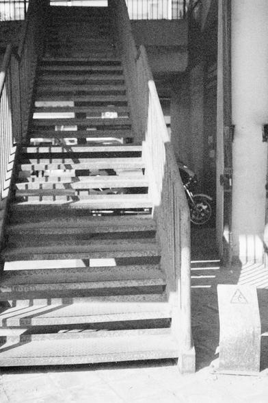 Ilford HP5 Plus 400 (FF)

English Alt Text: A black and white photo of a metal staircase with open risers, allowing light to cast shadows below. Railings run along both sides. To the right, a motorcycle is partially visible, parked in a shaded area. In the foreground, a concrete structure bears a triangular warning sign. The interplay of light and shadow adds drama to the industrial setting, emphasizing texture and geometry.

中文替代文字：
这是一张黑白照片，描绘了一座金属楼梯，楼梯踏板之间留有空隙，使光线能够穿透并在地面投下阴影。楼梯两侧设有栏杆。右侧阴影处停…