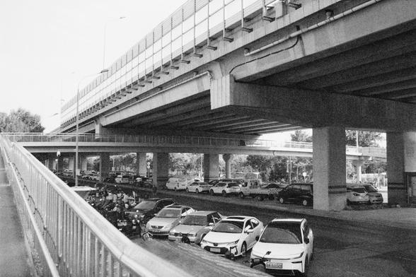 Ilford HP5 Plus 400 (FF)

English Alt Text: A black and white photo of a complex urban scene featuring multiple elevated highways crossing above a street-level area. Beneath the overpasses, cars and motorcycles are parked in a lot. A pedestrian walkway with railings runs along the left side, leading upward toward the elevated structures. The concrete pillars and layered roads dominate the composition, creating a sense of scale and depth. The monochrome palette emphasizes the contrast between th…