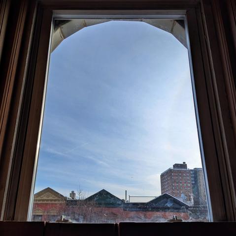 Looking through an arched window two hours after sunrise the blue sky is filled with diffuse streaky white clouds. Pointed roofs of Harlem brownstones with red brickwork are across the street, and a taller apartment building can be seen in the distance. The leafless tops of two trees are on the bottom and right. The window is grimy on the bottom.