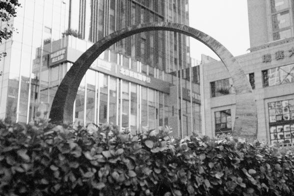 Ilford HP5 Plus 400 (FF)

English Alt Text: A black-and-white photo depicts an urban scene with a large semi-circular metal sculpture in the foreground, partially hidden by a hedge. Behind it are modern high-rise buildings with glass facades and Chinese signage. The buildings feature vertical lines and grid patterns, suggesting a commercial or business district. The contrast between the natural hedge and sleek architecture highlights the blend of nature and urban design.

中文替代文字： 这是一张黑白城市景观照片，前…