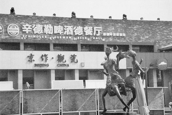 Ilford HP5 Plus 400 (FF)

English Alt Text: A black-and-white photo shows the exterior of Schindler German Beer Hall in Beijing. The signage includes both English and Chinese text, with logos for German beer brands like Weihenstephan above the entrance. Below, another sign reads “BEIJING CHINA.” In front of the building, a sculpture of two human figures in motion—possibly dancers—adds an artistic touch. The architecture blends German beer hall aesthetics with local Chinese elements, creating a …