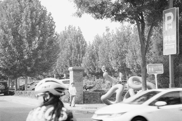 Ilford HP5 Plus 400 (FF)

English Alt Text: A black-and-white street scene captures a cyclist in the foreground wearing a helmet and patterned clothing. A white car partially obscures the view. In the background, tall trees line the street, and a person walks on the sidewalk. A large abstract sculpture with looping forms stands near the trees. Two signs are visible: one indicates a parking lot 50 meters ahead, and the other points to a public toilet. The image reflects a typical urban environme…